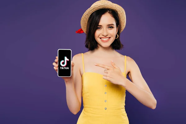 KYIV, UCRAINA - 3 LUGLIO 2019: ragazza sorridente con cappello di paglia puntata con il dito sullo smartphone con app tiktok isolata su viola — Foto stock