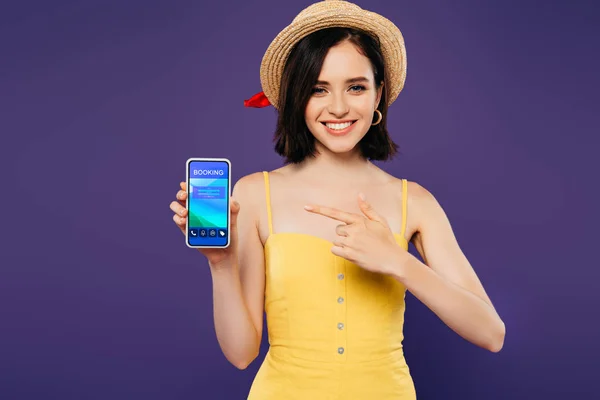 Chica sonriente en sombrero de paja apuntando con el dedo en el teléfono inteligente con aplicación de reserva aislado en púrpura - foto de stock