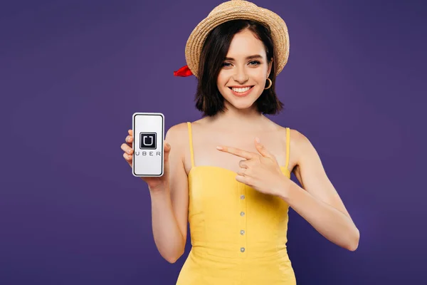 KYIV, UKRAINE - JULY 3, 2019: smiling girl in straw hat pointing with finger at smartphone with uber app isolated on purple — Stock Photo