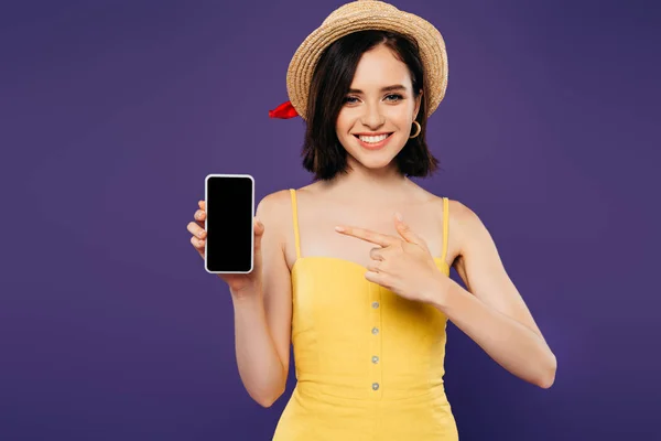 Улыбающаяся девушка в соломенной шляпе, указывающая пальцем на смартфон с пустым экраном, изолированным на фиолетовый — стоковое фото