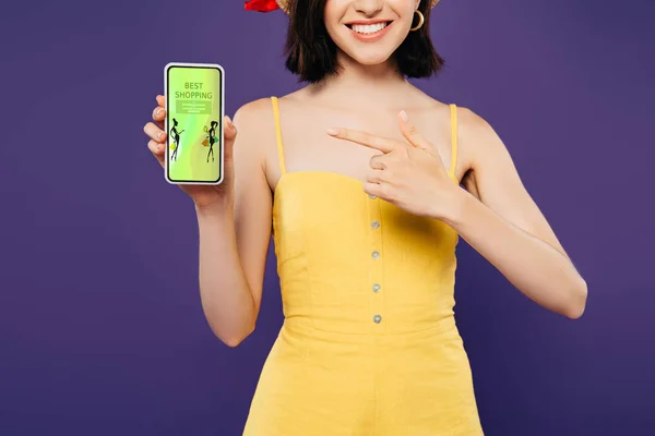 Обрезанный вид девушки в соломенной шляпе указывая пальцем на смартфон с лучшим приложением для покупок изолированы на фиолетовый — стоковое фото