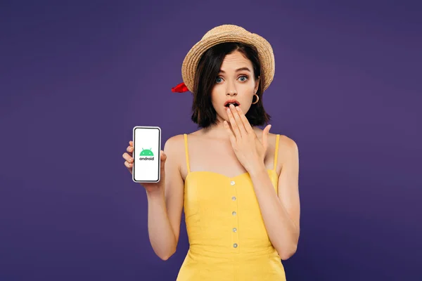 КИЕВ, Украина - 3 июля 2019 года: девушка в соломенной шляпе, показывающая жест идеи и держащая смартфон с логотипом андроида, изолированным на фиолетовом — стоковое фото