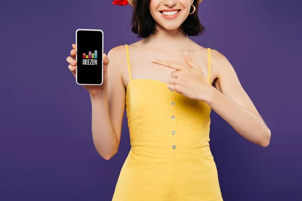 КИЕВ, Украина - 3 июля 2019 года: обрезанный вид девушки в соломенной шляпе, указывающей пальцем на смартфон с лиловым приложением, изолированным на фиолетовый — стоковое фото