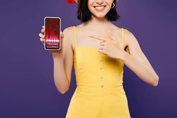 Vista cortada de menina em chapéu de palha apontando com o dedo para o smartphone com cursos comerciais aplicativo isolado em roxo — Fotografia de Stock