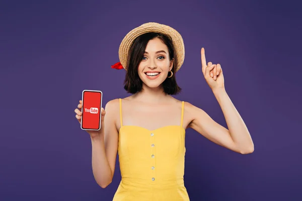 KYIV, UKRAINE - JULHO 3, 2019: garota animada de chapéu de palha mostrando gesto ideia e segurando smartphone com aplicativo youtube isolado em roxo — Fotografia de Stock