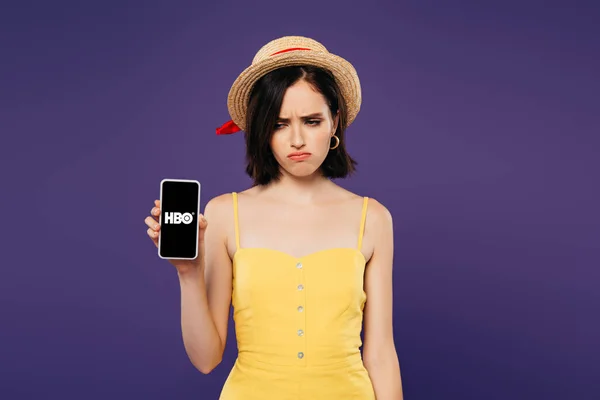 KYIV, UCRANIA - 3 de julio de 2019: triste chica bonita con sombrero de paja sosteniendo el teléfono inteligente con la aplicación HBO aislada en púrpura - foto de stock