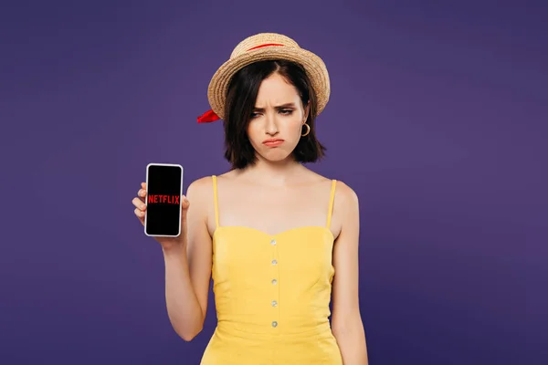 KYIV, UCRANIA - 3 de julio de 2019: triste chica bonita con sombrero de paja que sostiene el teléfono inteligente con la aplicación netflix aislada en púrpura - foto de stock