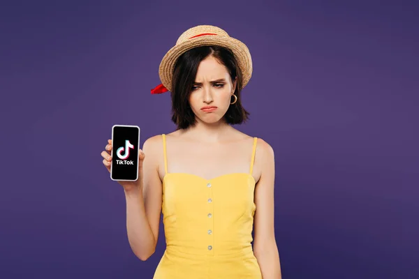 KYIV, UCRANIA - 3 de julio de 2019: triste chica bonita con sombrero de paja sosteniendo teléfono inteligente con aplicación tiktok aislado en púrpura - foto de stock