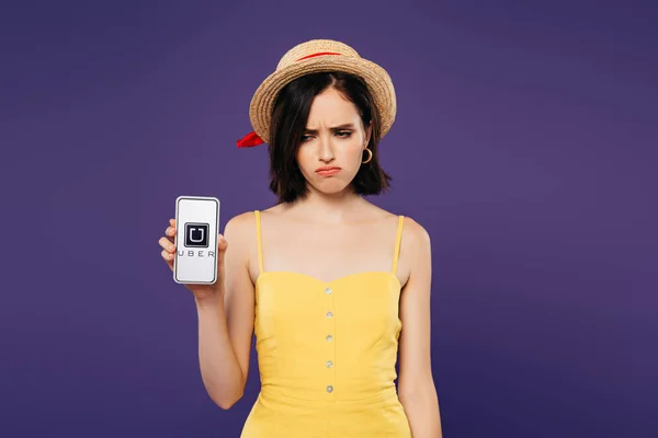 KYIV, UCRANIA - 3 de julio de 2019: triste chica bonita con sombrero de paja sosteniendo teléfono inteligente con aplicación uber aislado en púrpura - foto de stock