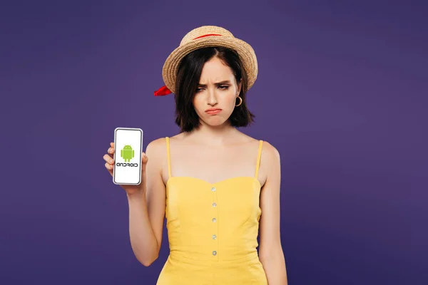 KYIV, UCRANIA - 3 de julio de 2019: triste chica bonita con sombrero de paja que sostiene el teléfono inteligente con el logotipo de Android aislado en púrpura - foto de stock