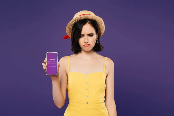 KYIV, UCRANIA - 3 de julio de 2019: triste chica bonita con sombrero de paja sosteniendo teléfono inteligente con aplicación instagram aislado en púrpura - foto de stock
