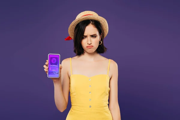 Triste chica bonita en sombrero de paja celebración de teléfono inteligente con aplicación de compras en línea aislado en púrpura - foto de stock