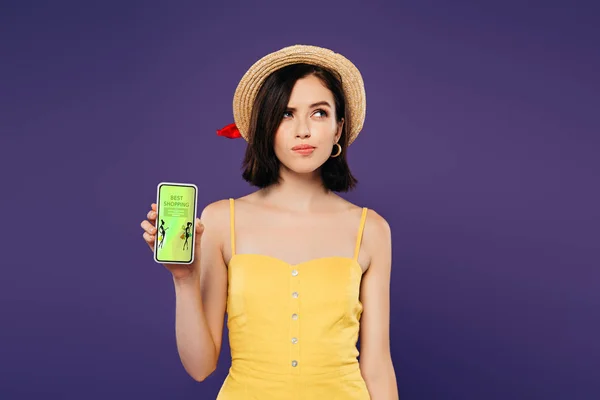 Sonhador menina bonita em chapéu de palha segurando smartphone com melhor aplicativo de compras isolado em roxo — Fotografia de Stock