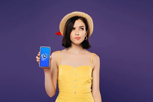 KYIV, UKRAINE - 3 JUILLET 2019 : jolie fille rêveuse en chapeau de paille tenant smartphone avec application shazam isolé sur violet — Photo de stock