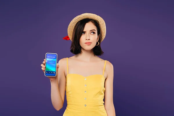 Verträumtes hübsches Mädchen mit Strohhut hält Smartphone mit Buchungs-App isoliert auf lila — Stockfoto