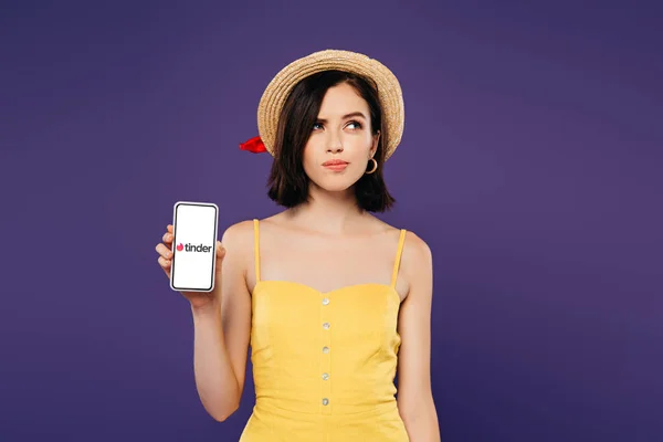 KYIV, UKRAINE - JULY 3, 2019: dreamy pretty girl in straw hat holding smartphone with tinder app isolated on purple — Stock Photo