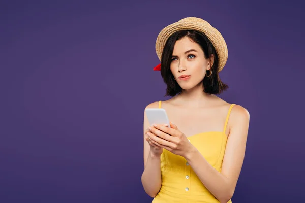 Jolie fille réfléchie en chapeau de paille en utilisant smartphone isolé sur violet — Photo de stock