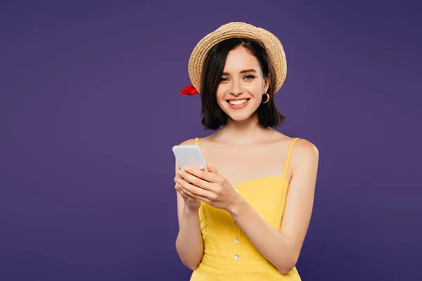 Sonriente chica bonita en sombrero de paja usando teléfono inteligente aislado en púrpura - foto de stock