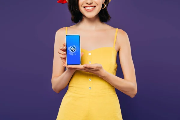 KYIV, UCRANIA - 3 de julio de 2019: vista recortada de una chica sonriente sosteniendo un teléfono inteligente con aplicación shazam aislada en púrpura - foto de stock