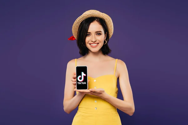 KYIV, UKRAINE - 3 JUILLET 2019 : jolie fille souriante en chapeau de paille tenant smartphone avec application tiktok isolée sur violet — Photo de stock