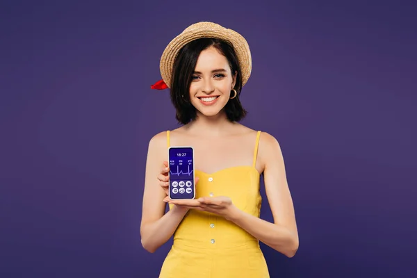 Sonriente chica bonita en sombrero de paja celebración de teléfono inteligente con aplicación sanitaria aislado en púrpura - foto de stock