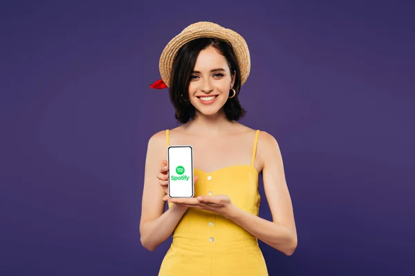 KYIV, UKRAINE - 3 JUILLET 2019 : jolie fille souriante en chapeau de paille tenant smartphone avec application spotify isolé sur violet — Photo de stock