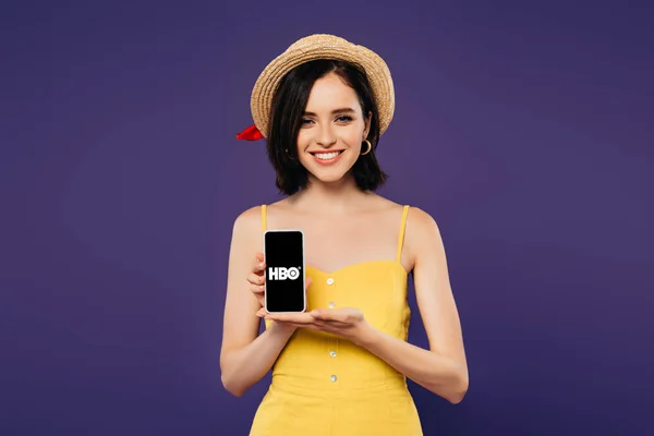 КИЕВ, УКРАИНА - 3 июля 2019 года: улыбающаяся красивая девушка в соломенной шляпе держит смартфон с приложением HBO изолированным на фиолетовый — стоковое фото