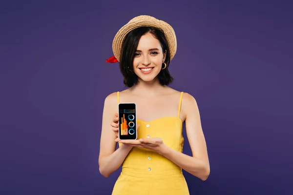 Sorrindo menina bonita em chapéu de palha segurando smartphone com aplicativo de negócios isolado em roxo — Fotografia de Stock