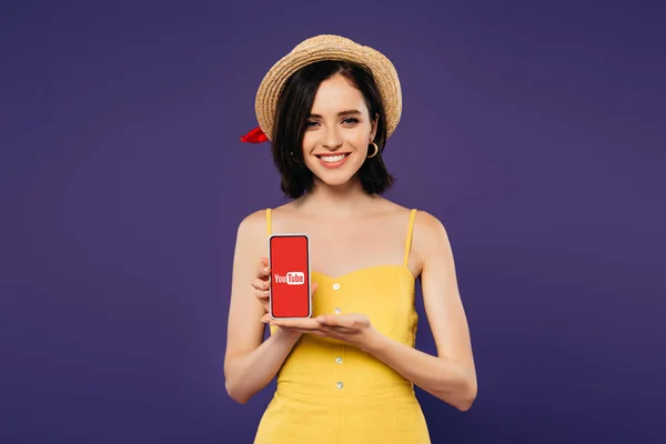 KYIV, UCRAINA - 3 LUGLIO 2019: bella ragazza sorridente in cappello di paglia con smartphone in mano con app youtube isolata su viola — Foto stock
