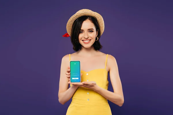 KYIV, UKRAINE - 3 JUILLET 2019 : jolie fille souriante en chapeau de paille tenant smartphone avec application twitter isolée sur violet — Photo de stock