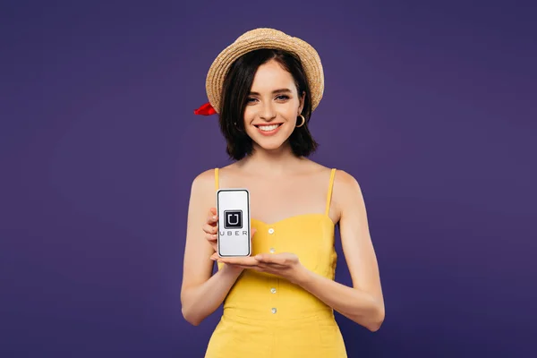 KYIV, UCRAINA - LUGLIO 3, 2019: bella ragazza sorridente in cappello di paglia con smartphone in mano con app uber isolata su viola — Foto stock