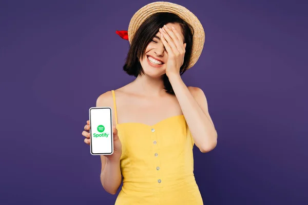 Київ, Україна-3 липня 2019: усміхнена красива дівчина в солом'яному капелюсі з рукою на обличчі показує смартфон з додатком Spotify, ізольованим від фіолетового — стокове фото