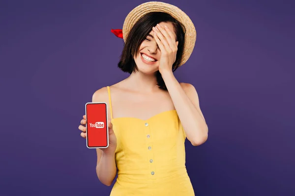 KYIV, UCRANIA - JULIO 3, 2019: sonriente chica bonita en sombrero de paja con la mano en la cara que muestra el teléfono inteligente con aplicación de youtube aislado en púrpura - foto de stock