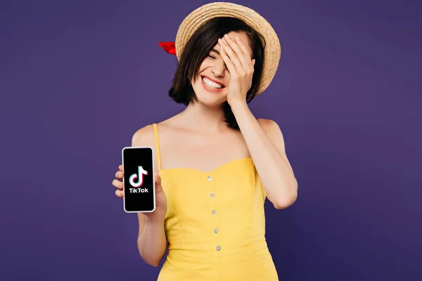 KYIV, UCRANIA - JULIO 3, 2019: sonriente chica bonita en sombrero de paja con la mano en la cara que muestra el teléfono inteligente con aplicación tiktok aislado en púrpura - foto de stock