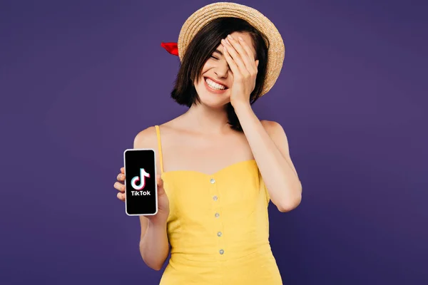 KYIV, UCRAINA - LUGLIO 3, 2019: bella ragazza sorridente con cappello di paglia e faccia a faccia che mostra smartphone con app tiktok isolata su viola — Foto stock