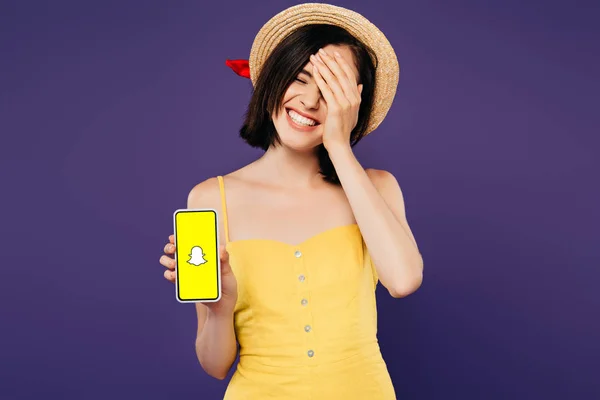 КИЕВ, УКРАИНА - 3 ИЮЛЯ 2019 года: улыбающаяся красивая девушка в соломенной шляпе с рукой на лице, показывающая смартфон с приложением Snapchat, изолированным на фиолетовый — стоковое фото