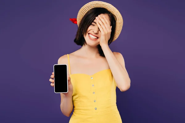 Sorridente bella ragazza in cappello di paglia con mano sul viso mostrando smartphone con schermo bianco isolato su viola — Foto stock