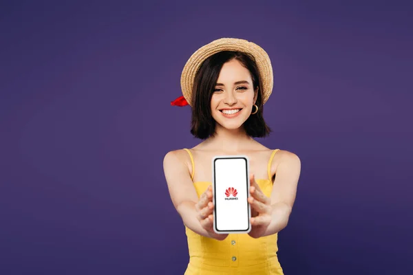 KYIV, UKRAINE - 3 JUILLET 2019 : mise au point sélective d'une jolie fille souriante en chapeau de paille présentant un smartphone avec logo géant isolé sur violet — Photo de stock