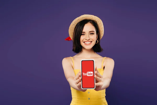 KYIV, UKRAINE - JULY 3, 2019: selective focus of smiling pretty girl in straw hat presenting smartphone with youtube app isolated on purple — Stock Photo