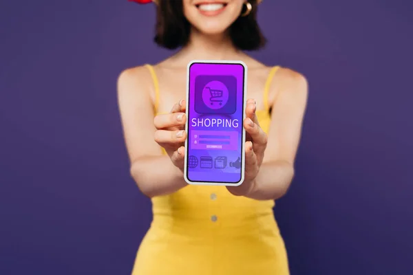 Enfoque selectivo de chica sonriente presentando teléfono inteligente con aplicación de compras en línea aislado en púrpura - foto de stock