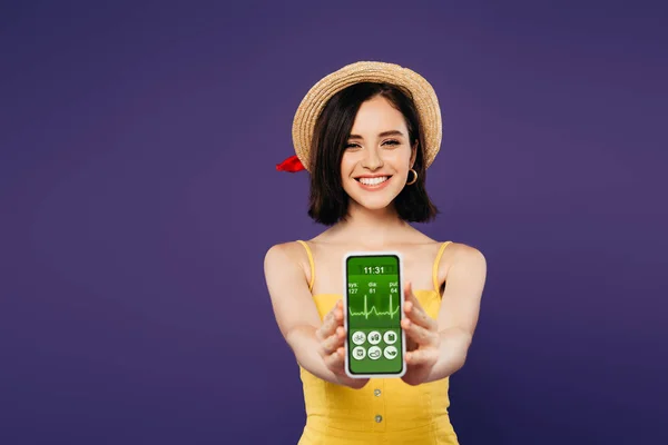 Селективный фокус улыбающейся красивой девушки в соломенной шляпе, представляющей смартфон с приложением для здравоохранения, изолированным на фиолетовый — стоковое фото