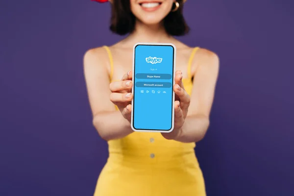 КИЕВ, УКРАИНА - 3 июля 2019 года: избирательный фокус улыбающейся девушки, представляющей смартфон с приложением skype, изолированным на фиолетовый — стоковое фото