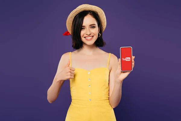 KYIV, UKRAINE - JULY 3, 2019: smiling pretty girl in straw hat showing thumb up while holding smartphone with youtube app isolated on purple — Stock Photo