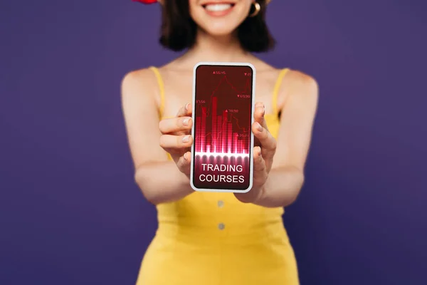 Selektiver Fokus des lächelnden Mädchens präsentiert Smartphone mit Trading Courses App isoliert auf lila — Stockfoto