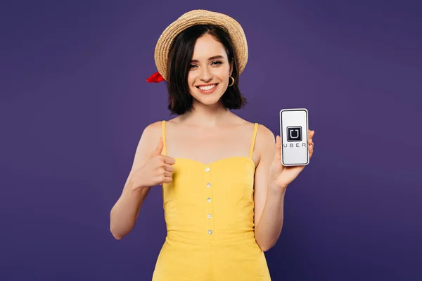 KYIV, UKRAINE - JULY 3, 2019: smiling pretty girl in straw hat showing thumb up while holding smartphone with uber app isolated on purple — Stock Photo