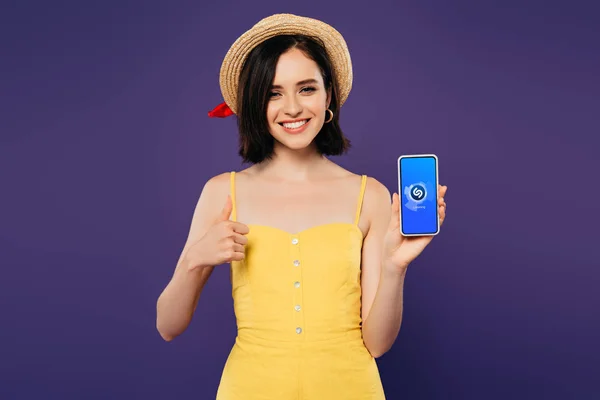 KYIV, UKRAINE - JULY 3, 2019: smiling pretty girl in straw hat showing thumb up while holding smartphone with shazam app isolated on purple — Stock Photo