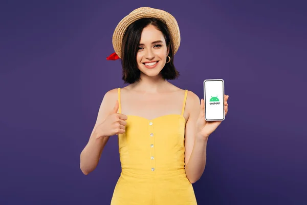 Київ, Україна-3 липня 2019: усміхнена красива дівчина в солом'яному капелюсі показує великий палець, утримуючи смартфон з логотипом Android, ізольованих на фіолетовий — стокове фото