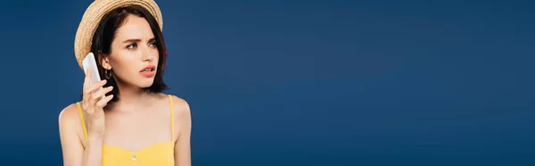 Панорамний знімок сумної красивої дівчини в солом'яному капелюсі, що говорить на смартфоні ізольовано на синьому — стокове фото