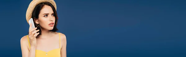 Panoramic shot of sad pretty girl in straw hat talking on smartphone isolated on blue — Stock Photo