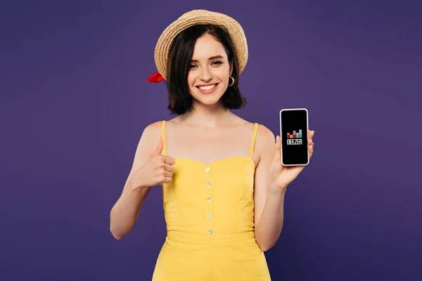 КИЕВ, УКРАИНА - 3 июля 2019 года: улыбающаяся красивая девушка в соломенной шляпе показывает большой палец вверх, держа смартфон с приложением Deezer изолированным на фиолетовый — стоковое фото